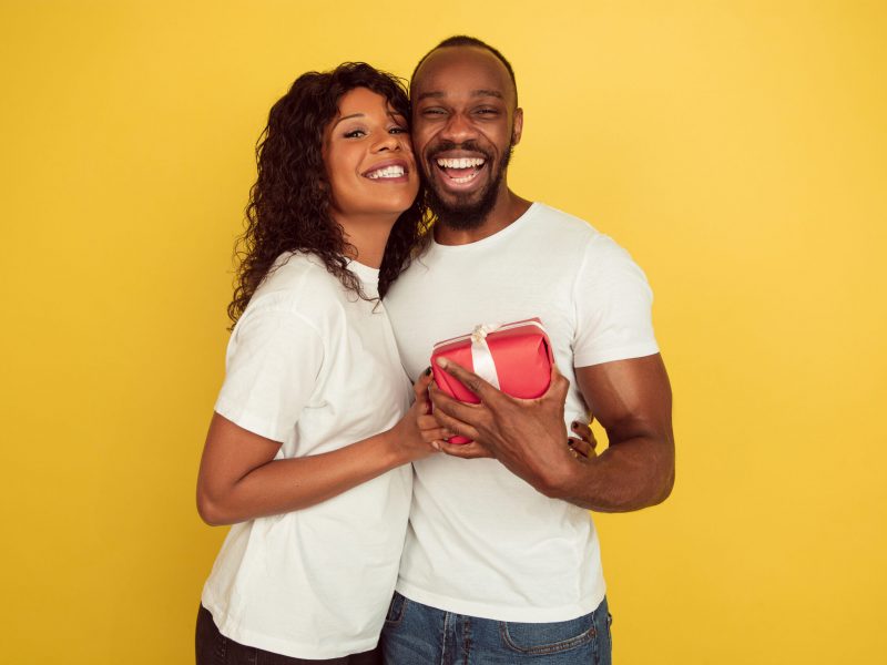 Giving surprise. Valentine's day celebration, happy african-american couple isolated on yellow studio background. Concept of human emotions, facial expression, love, relations, romantic holidays.