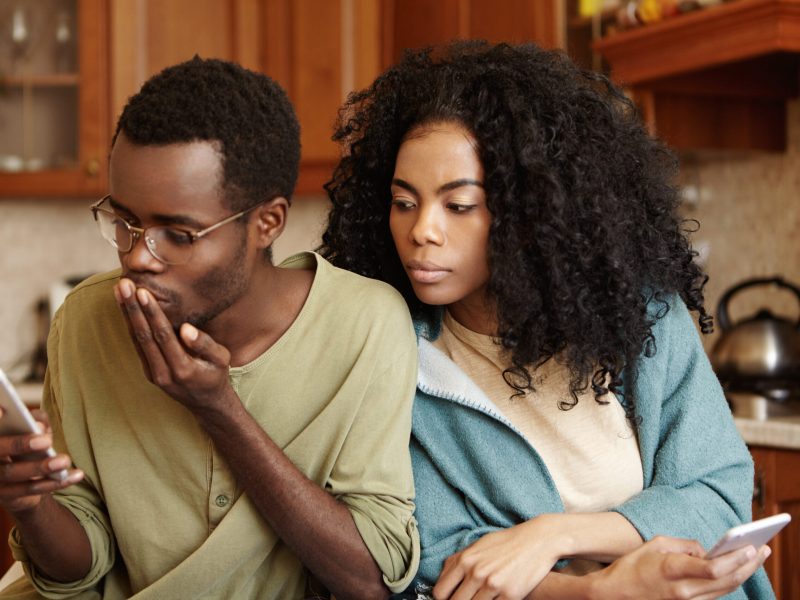 Handsome African-American male in glasses reading sms on mobile phone, messaging lover while his possessive beautiful wife spying on him, looking at screen of electronic gadget with sneaky expression