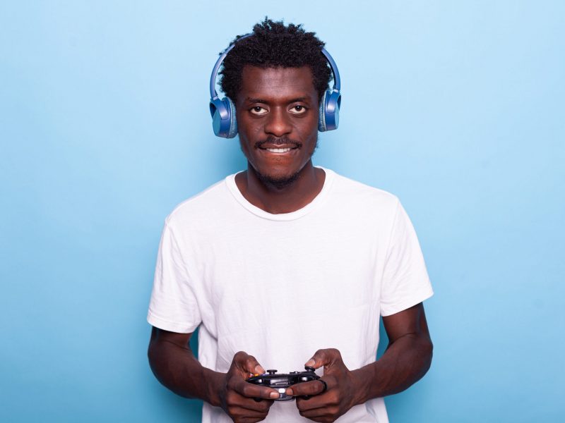 Portrait of man playing video games with controller while wearing headphones in studio. Person listening to music and holding joystick for game play on console, looking at camera